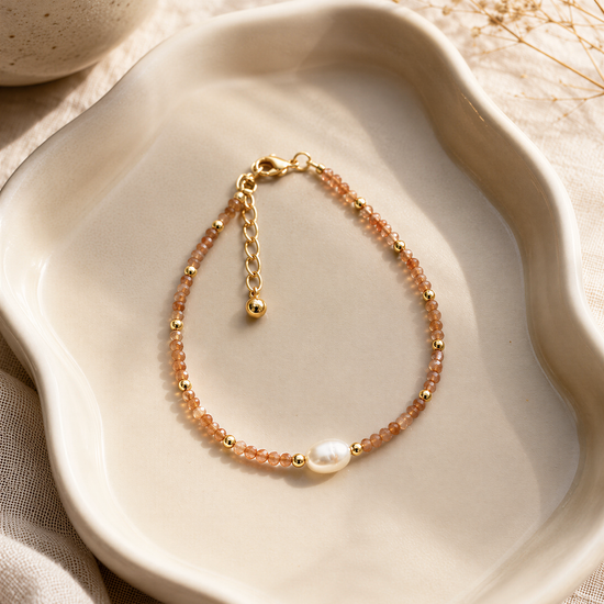 A delicate gold-colored bracelet with a pearl bead and beaded design rests on a white plate, accompanied by dried flowers and a white ceramic bowl.