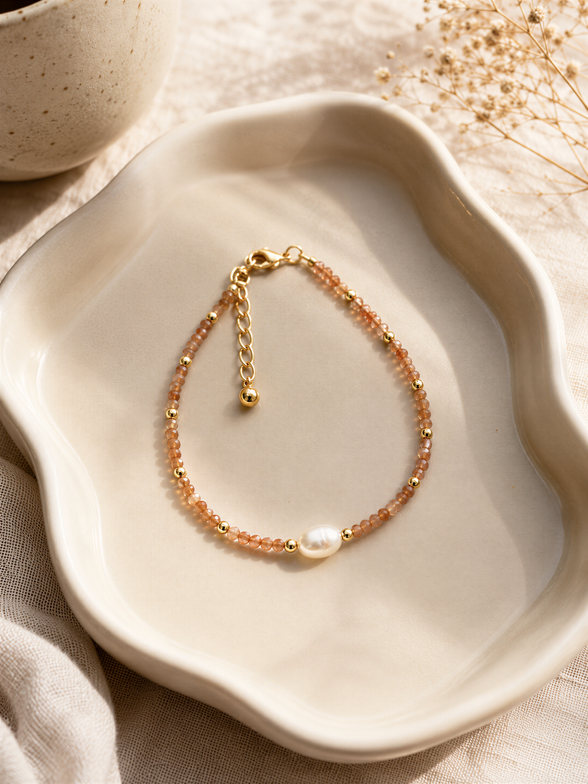 A delicate gold-colored bracelet with a pearl bead and beaded design rests on a white plate, accompanied by dried flowers and a white ceramic bowl.