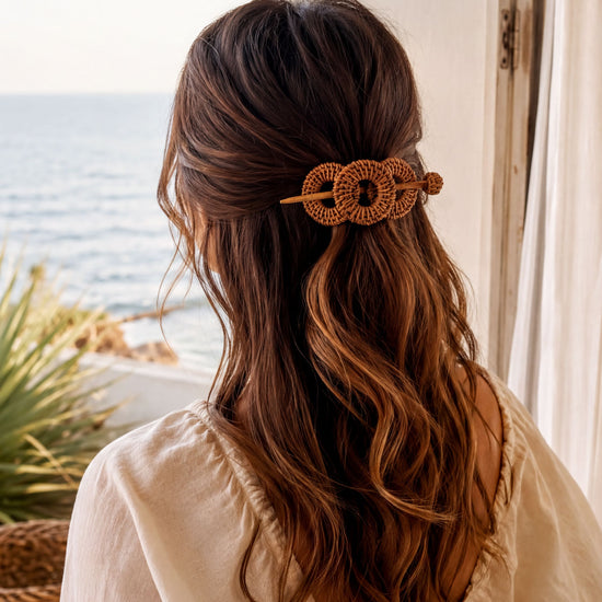 a woman with long, wavy brown hair wearing a white blouse and a hair accessory with a circular design. She is looking out a window, with the ocean visible in the background.