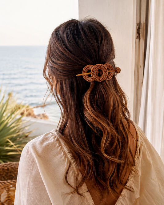 a woman with long, wavy brown hair wearing a white blouse and a hair accessory with a circular design. She is looking out a window, with the ocean visible in the background.