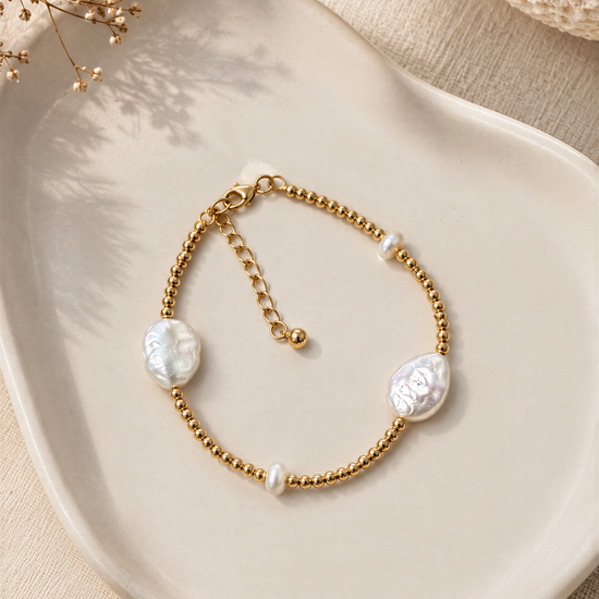 A gold-colored bracelet with pearl-like stones is displayed on a white plate, accompanied by dried flowers and a white fabric background.
