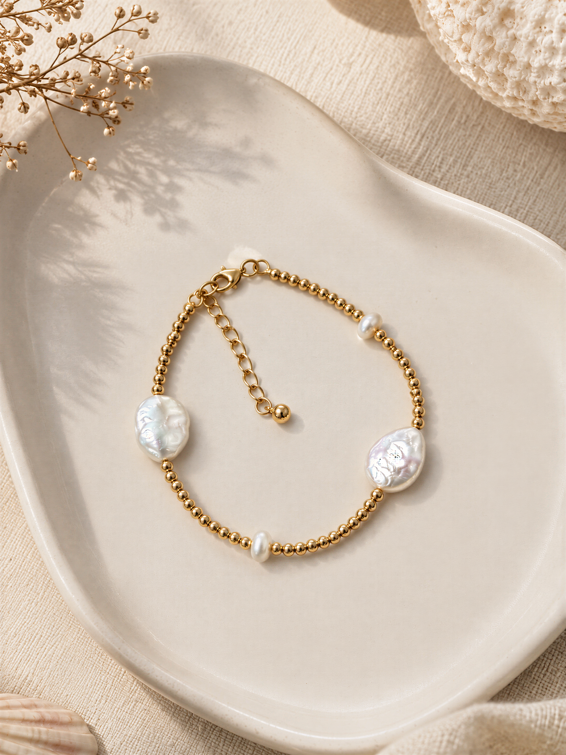 A gold-colored bracelet with pearl-like stones is displayed on a white plate, accompanied by dried flowers and a white fabric background.