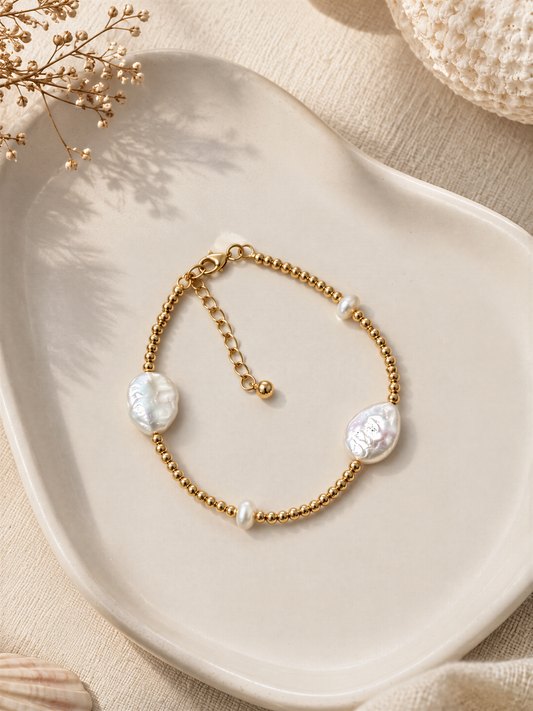 A gold-colored bracelet with pearl-like stones is displayed on a white plate, accompanied by dried flowers and a white fabric background.