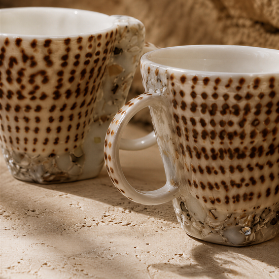 Two white mugs with brown speckled patterns are placed on a textured surface.
