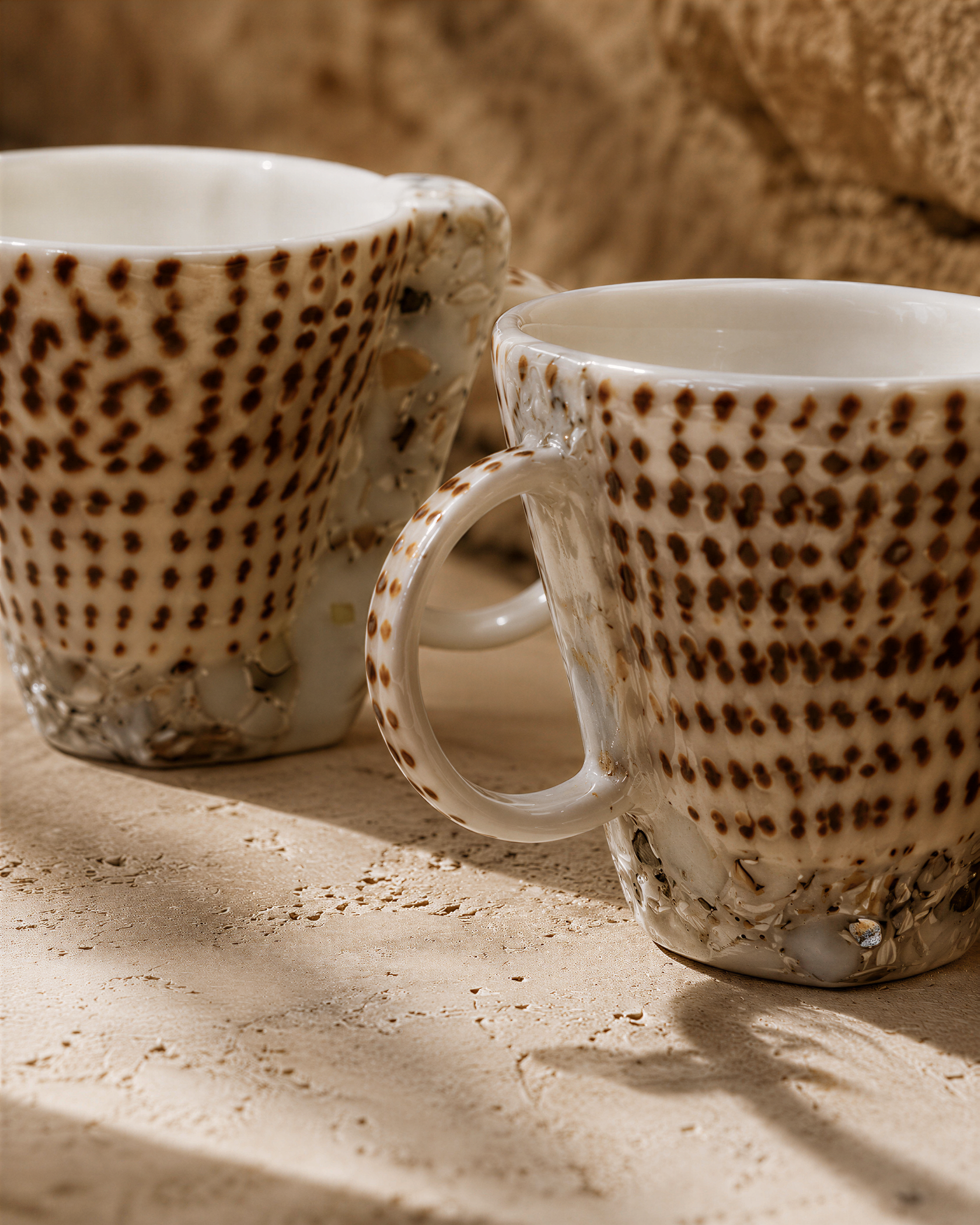 Two white mugs with brown speckled patterns are placed on a textured surface.