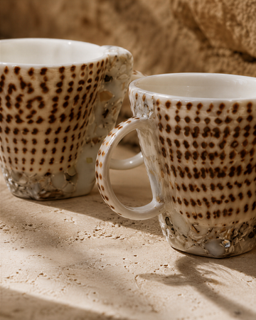 Two white mugs with brown speckled patterns are placed on a textured surface.