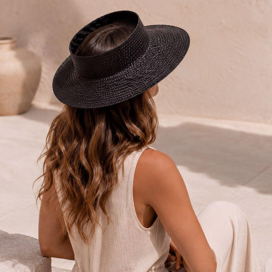 A woman wearing a black hat sits on a white surface, facing away from the camera.