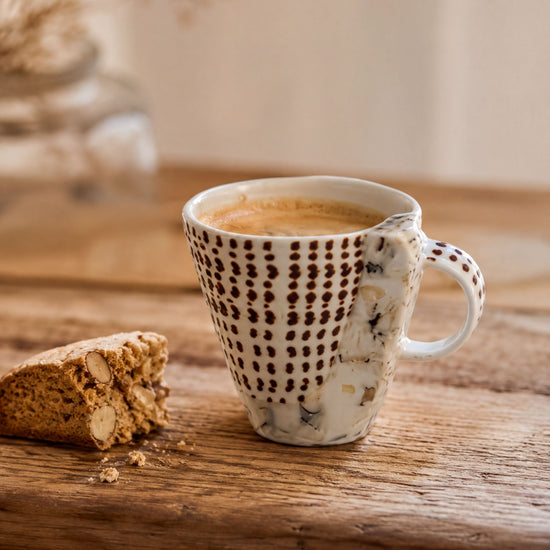 A white mug with a black and white polka dot pattern sits on a wooden surface, with a partially eaten cookie beside it.