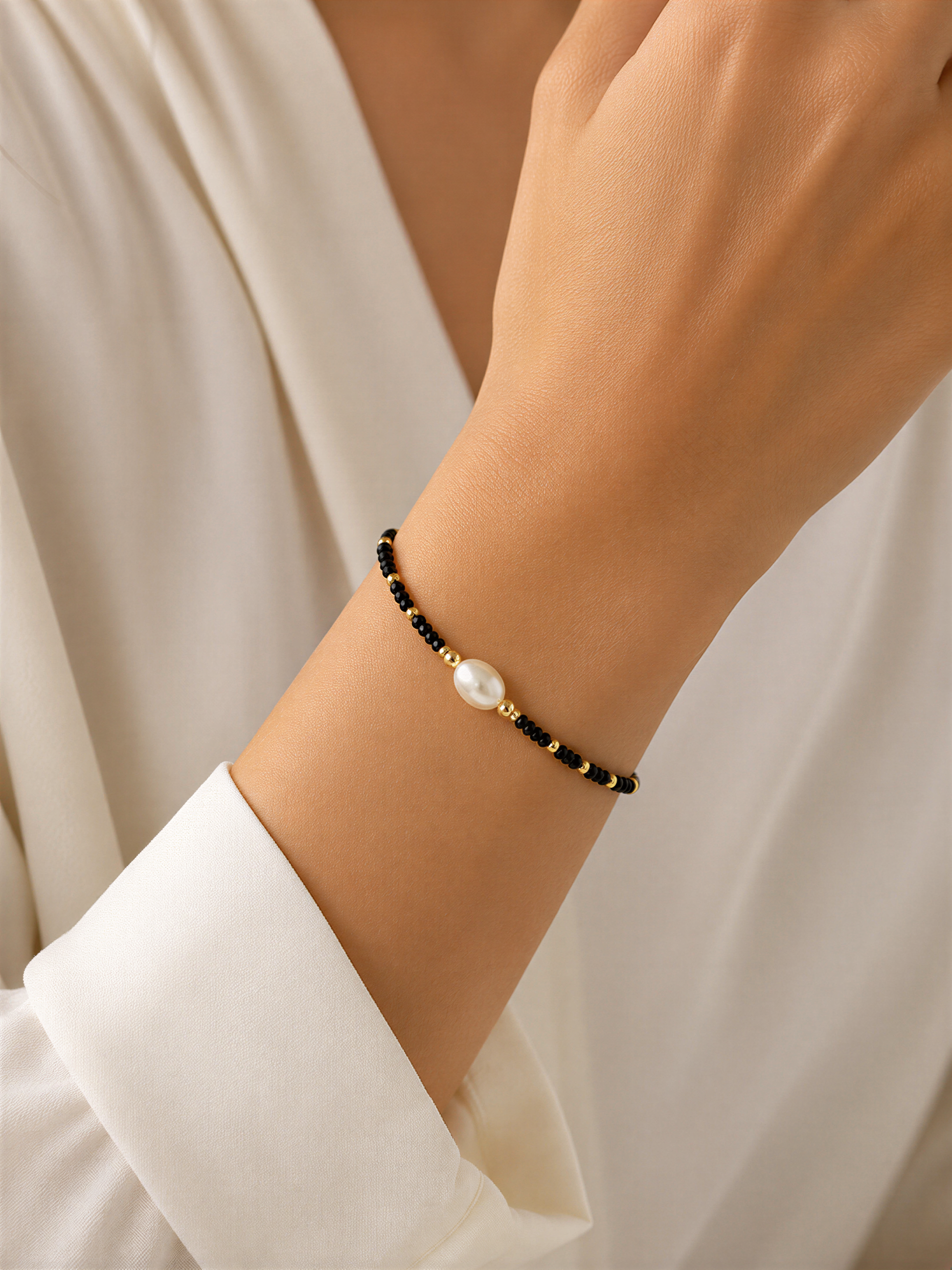 A close-up view of a person's wrist wearing a black and gold bead bracelet with a single pearl bead.