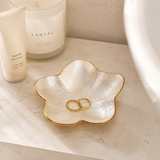 a white candle labeled "Santal" and a white tube of cream, both placed on a white surface. There is also a white bowl containing a gold ring and a white bowl containing a gold ring.