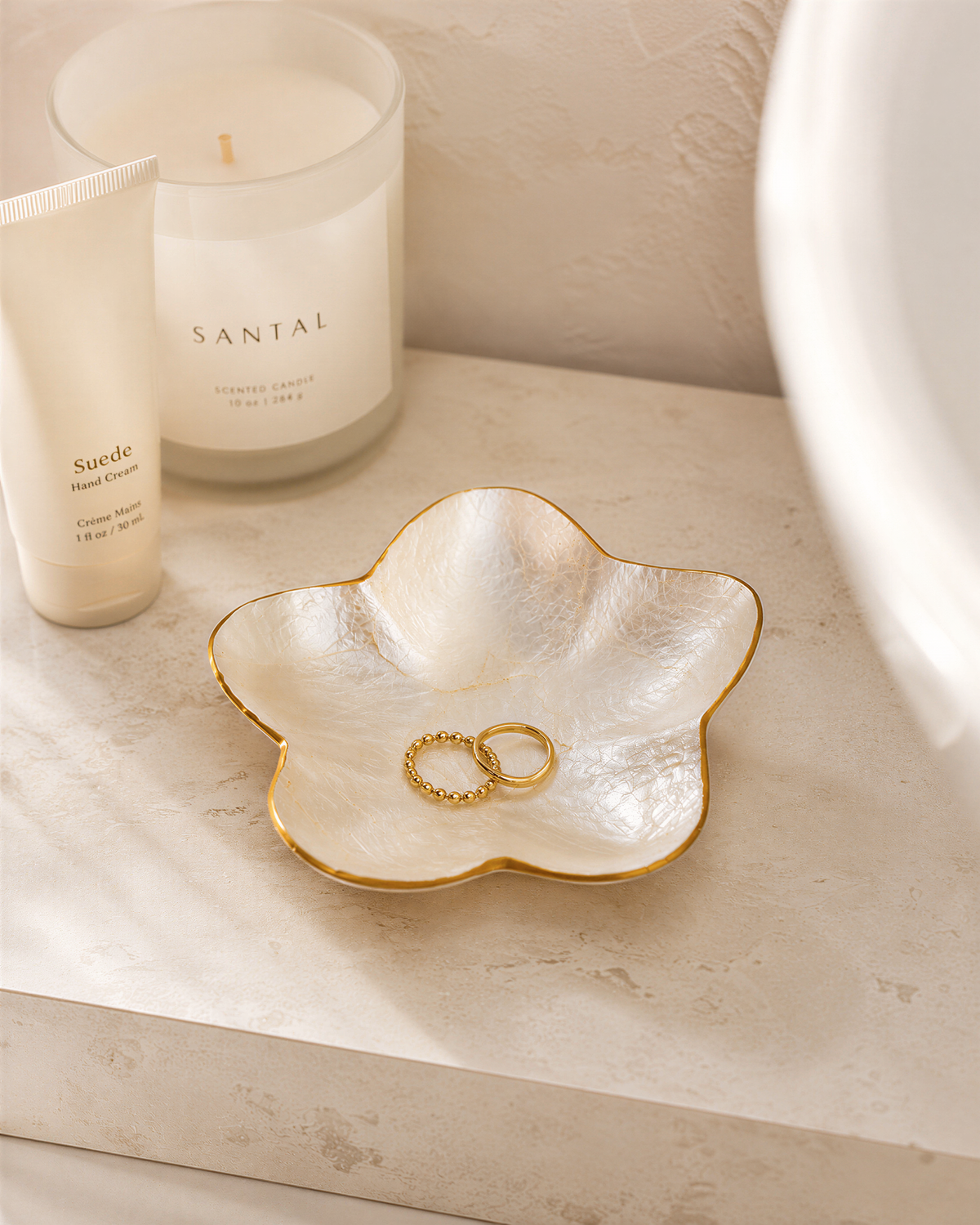 a white candle labeled "Santal" and a white tube of cream, both placed on a white surface. There is also a white bowl containing a gold ring and a white bowl containing a gold ring.