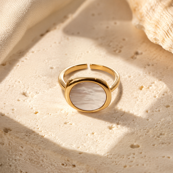 A gold ring with a white oval-shaped stone is displayed on a textured surface, possibly a piece of fabric or a white background.