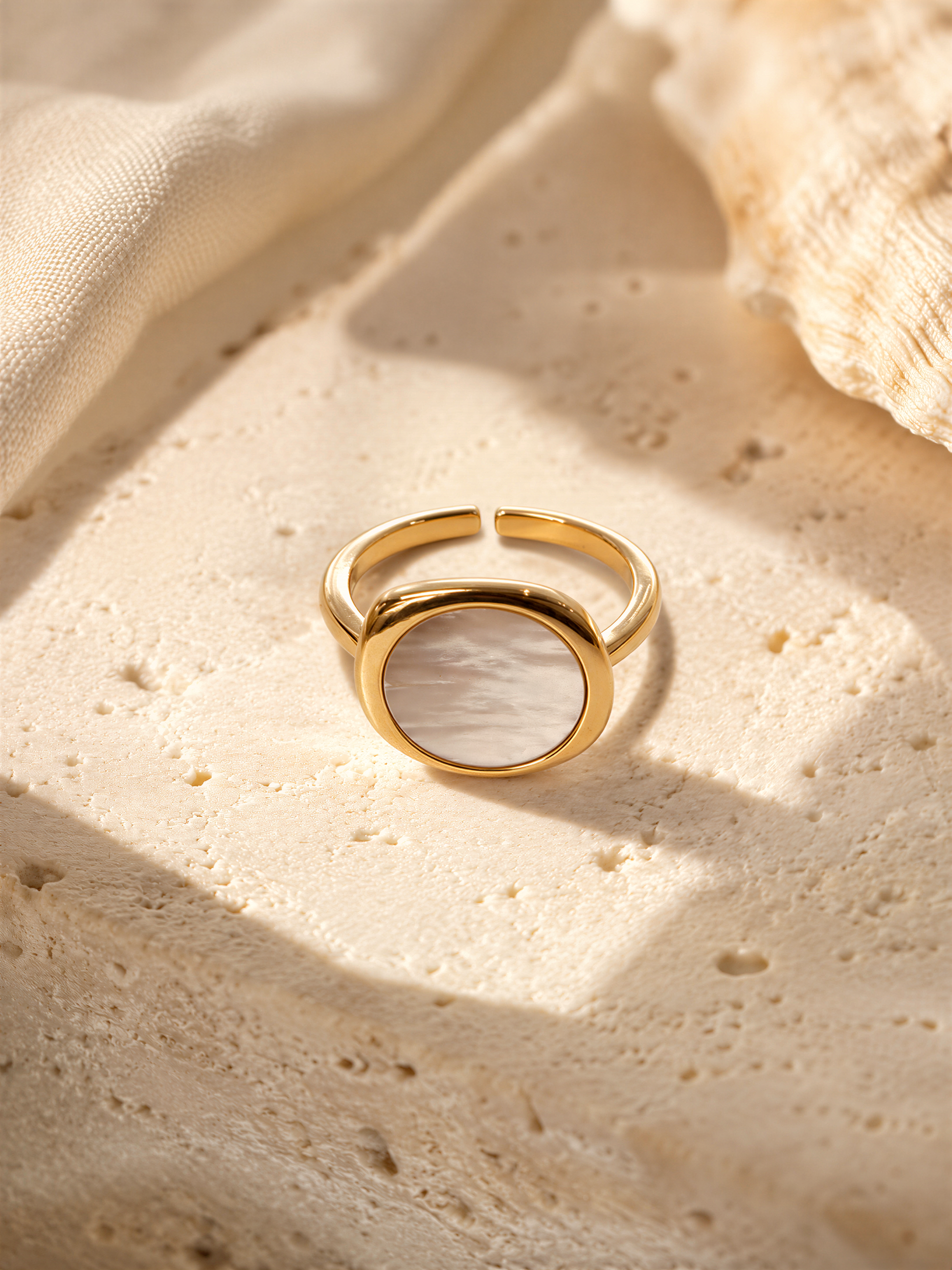 A gold ring with a white oval-shaped stone is displayed on a textured surface, possibly a piece of fabric or a white background.