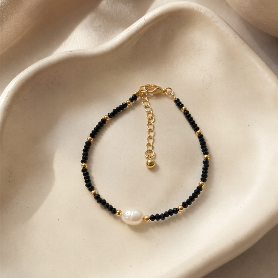 A black and gold beaded bracelet with a pearl accent is displayed on a white plate.