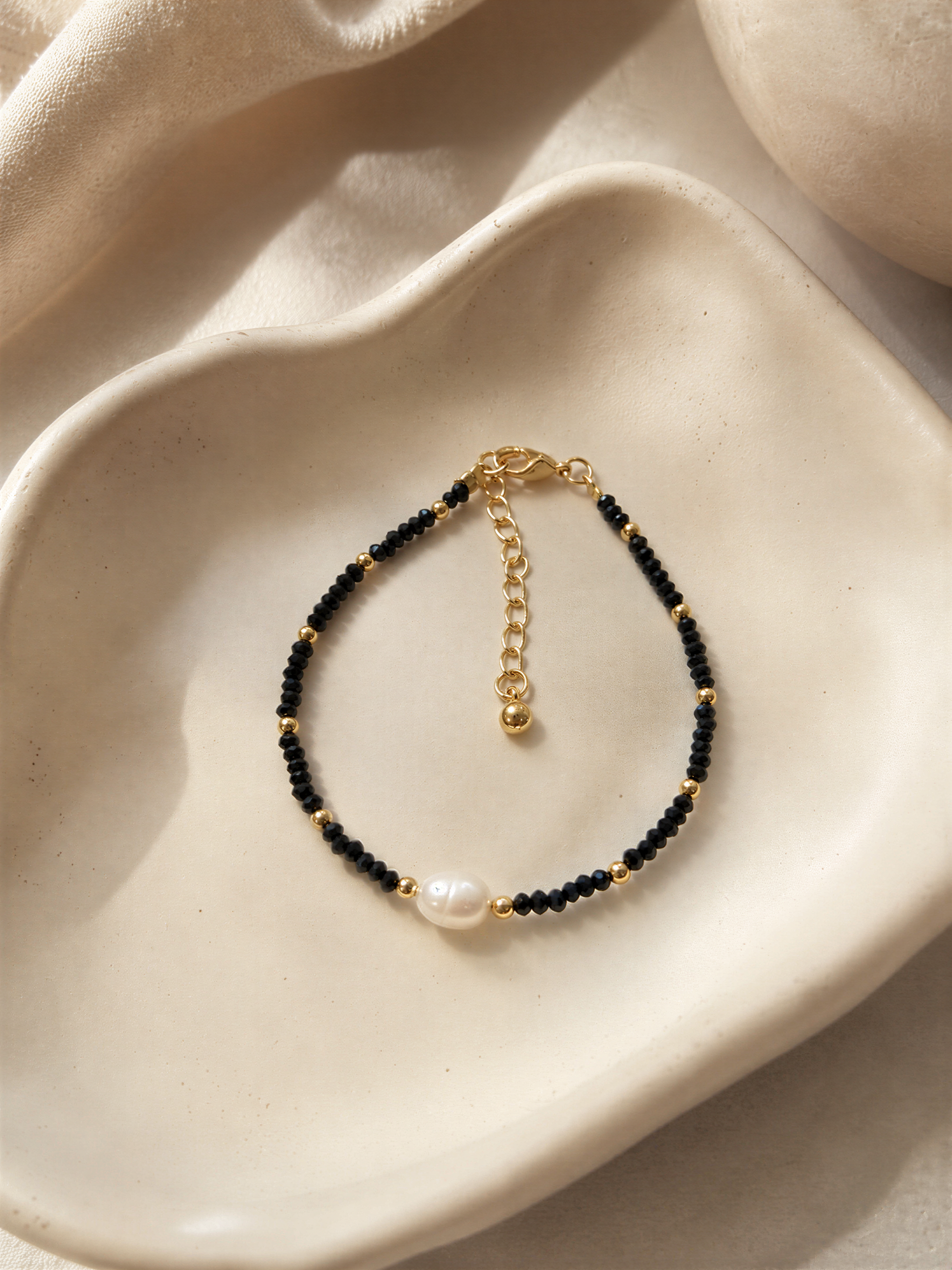 A black and gold beaded bracelet with a pearl accent is displayed on a white plate.