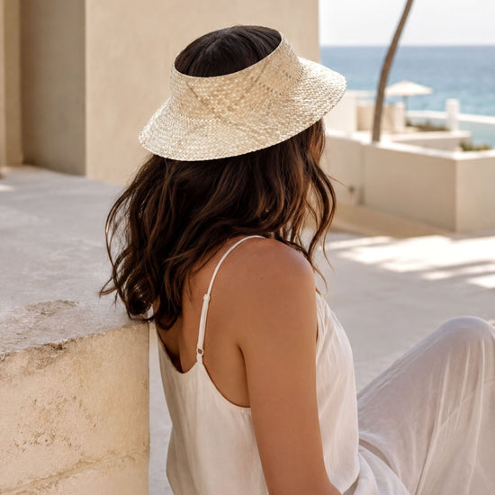 A woman in a white dress and straw hat sits on a stone wall, gazing out at the ocean.