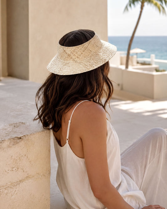 A woman in a white dress and straw hat sits on a stone wall, gazing out at the ocean.