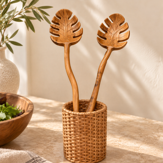 a wicker basket holding two wooden spoons with leaf-shaped handles, placed on a table with a bowl of greens and a vase of greenery in the background.