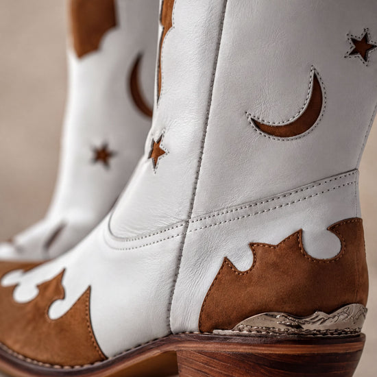 A close-up of a pair of white cowboy boots with brown accents, including star and crescent motifs, and a wooden heel.
