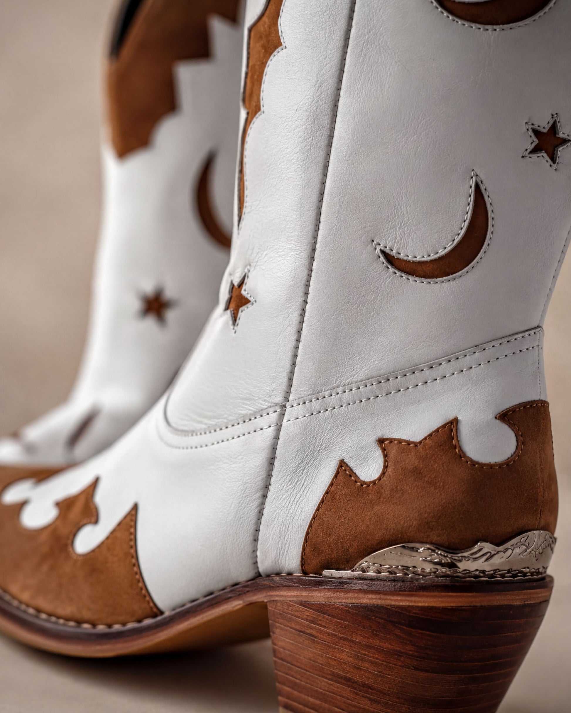 A close-up of a pair of white cowboy boots with brown accents, including star and crescent motifs, and a wooden heel.