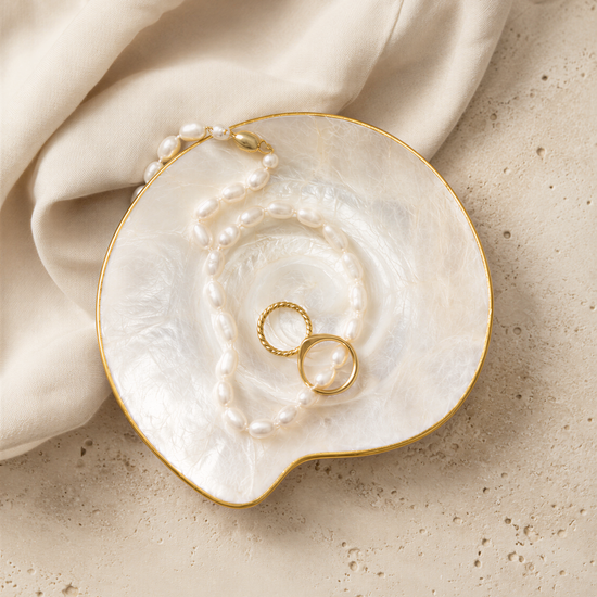 A white pearl necklace and two gold rings are displayed on a white, textured surface.
