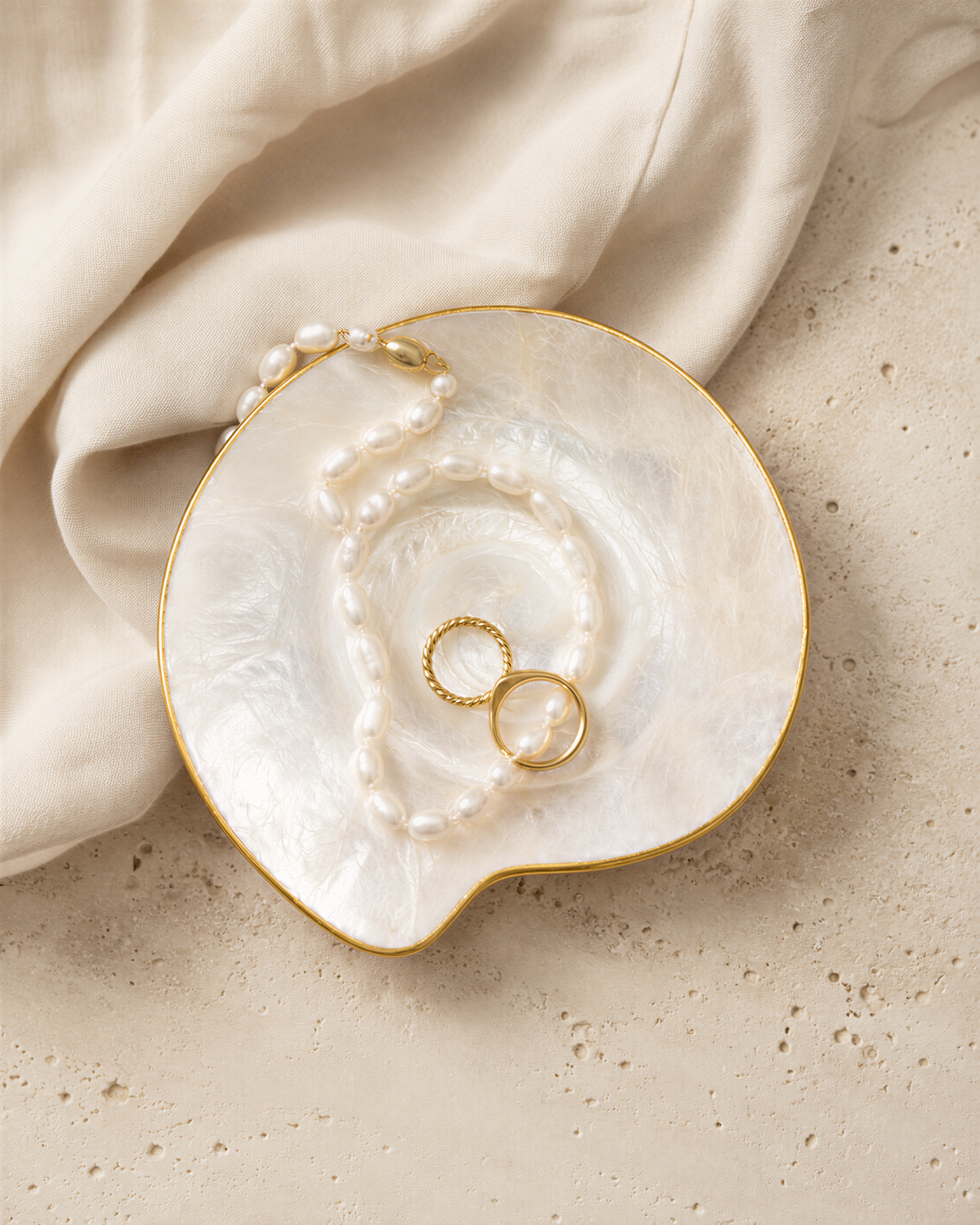 A white pearl necklace and two gold rings are displayed on a white, textured surface.